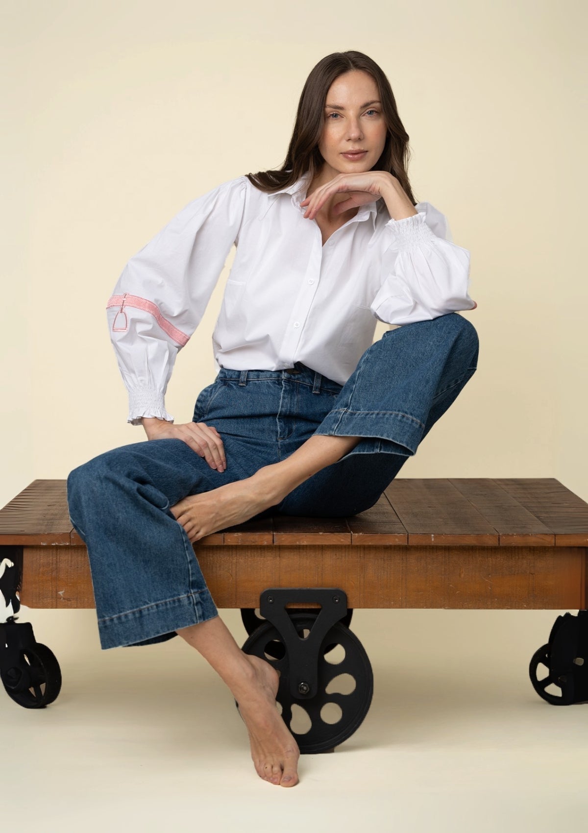 Mujer sentada en un banco de madera vistiendo una blusa blanca bordada y jeans azules sobre fondo beige.