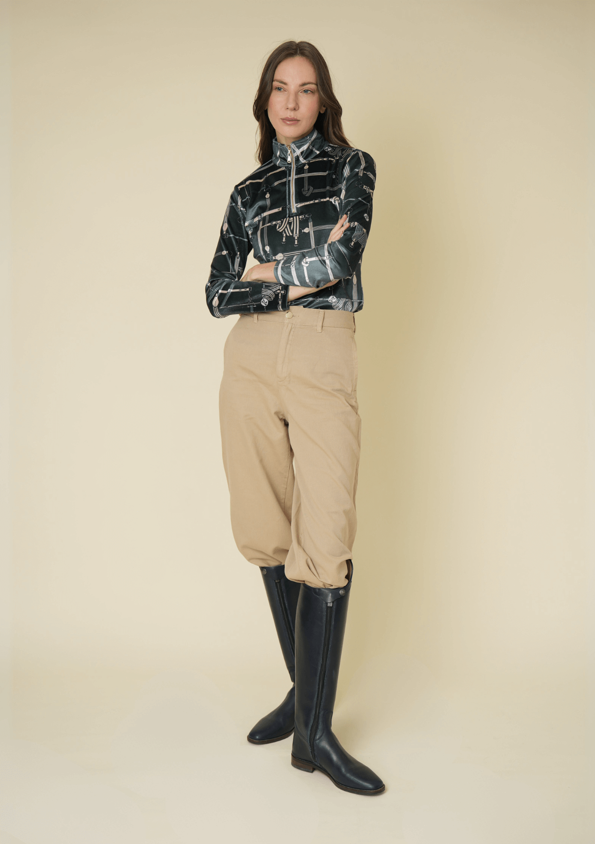 Woman wearing a patterned top, beige riding pants, and black riding boots on a beige background