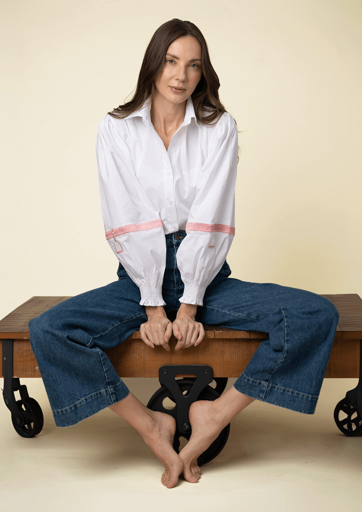 Mujer sentada en un banco de madera vistiendo una blusa blanca bordada y jeans azules sobre fondo beige.