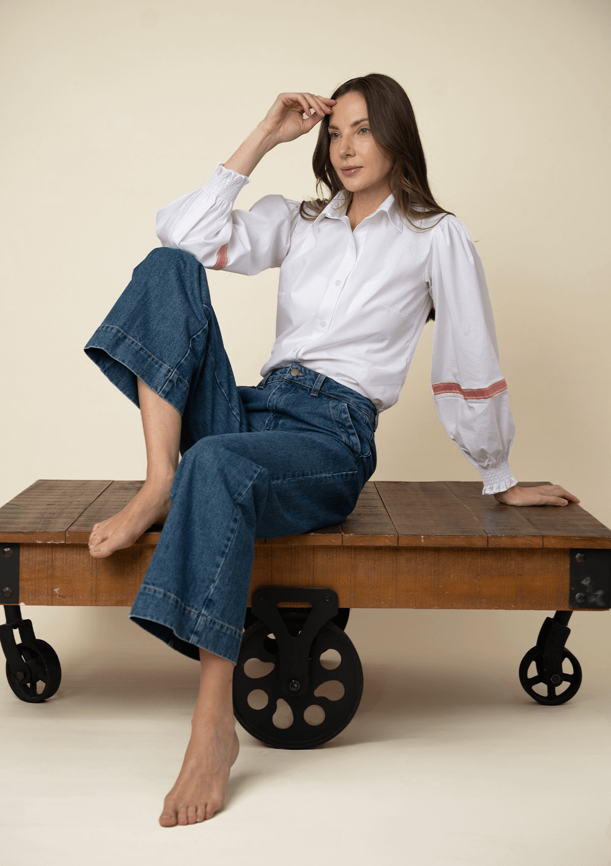 Mujer sentada en un banco de madera vistiendo una blusa blanca bordada y jeans azules sobre fondo beige.