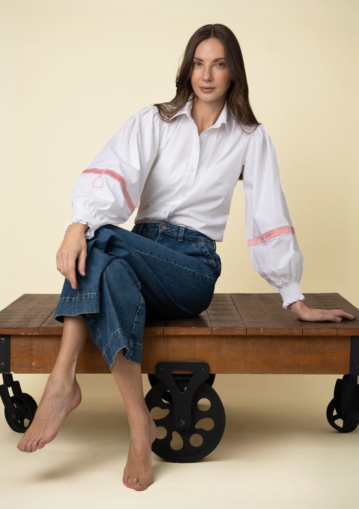 Mujer sentada en un banco de madera vistiendo una blusa blanca bordada y jeans azules sobre fondo beige.