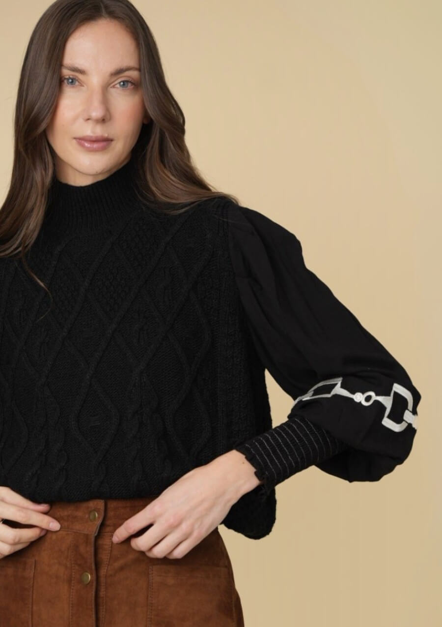 Mujer vistiendo un suéter negro de punto trenzado con detalles ecuestres tipo horsebit sobre fondo beige.

