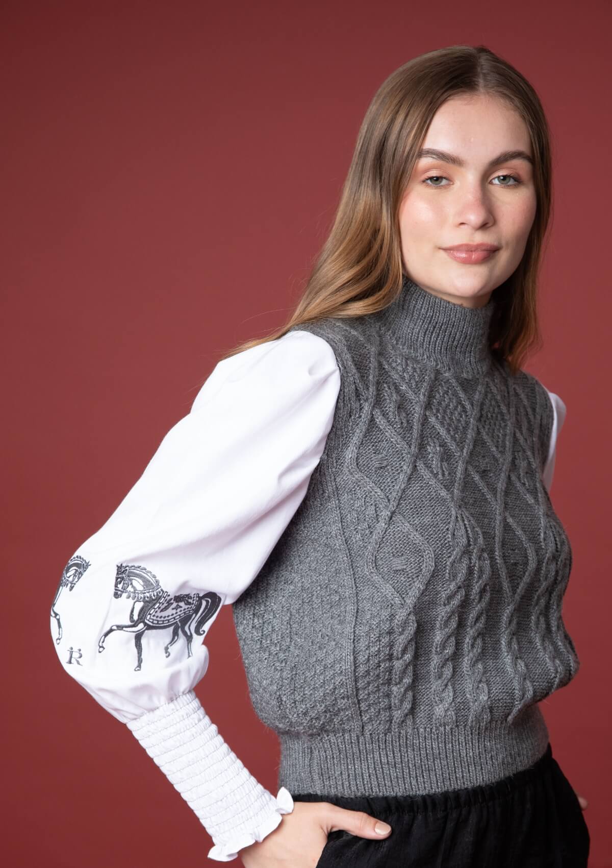 Mujer vistiendo un suéter gris de punto trenzado con detalles ecuestres tipo horsebit sobre fondo rojo.
