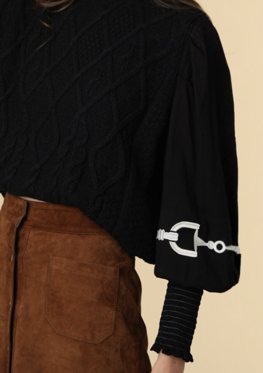 un suéter negro de punto trenzado con detalles ecuestres tipo horsebit sobre fondo beige.
