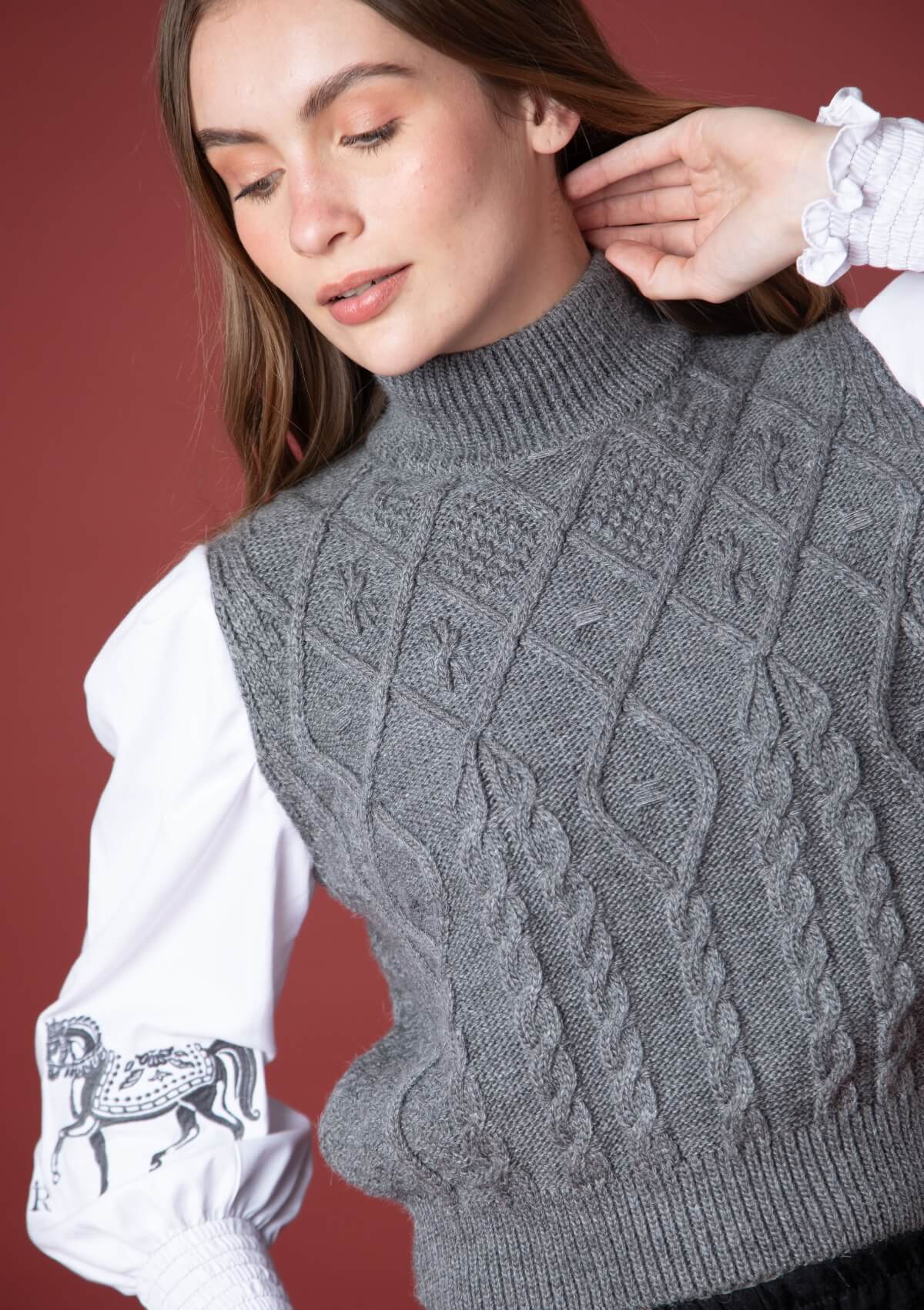 Mujer vistiendo un suéter gris de punto trenzado con detalles ecuestres tipo horsebit sobre fondo rojo.
