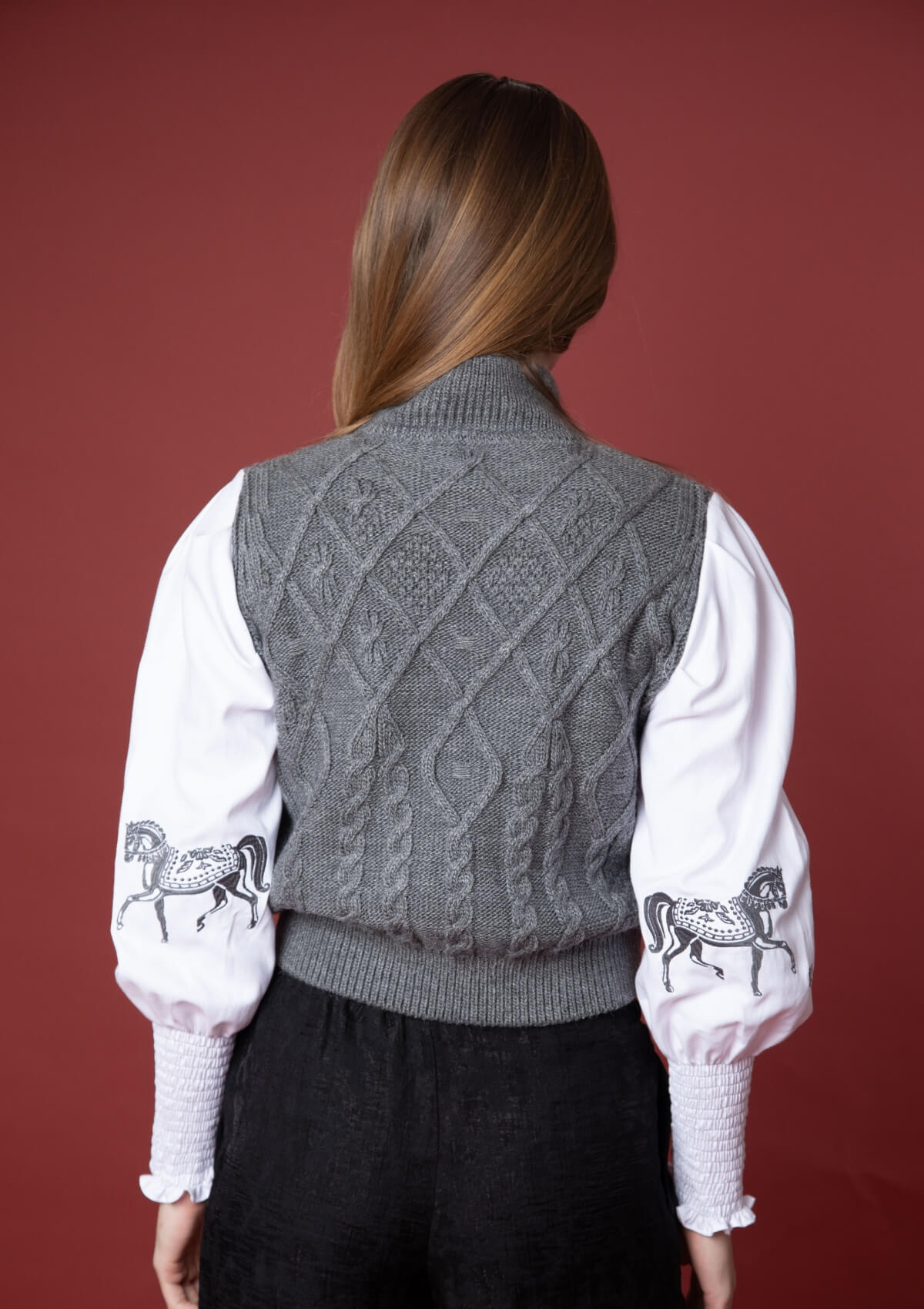 Mujer vistiendo un suéter gris de punto trenzado con detalles ecuestres tipo horsebit sobre fondo rojo.
